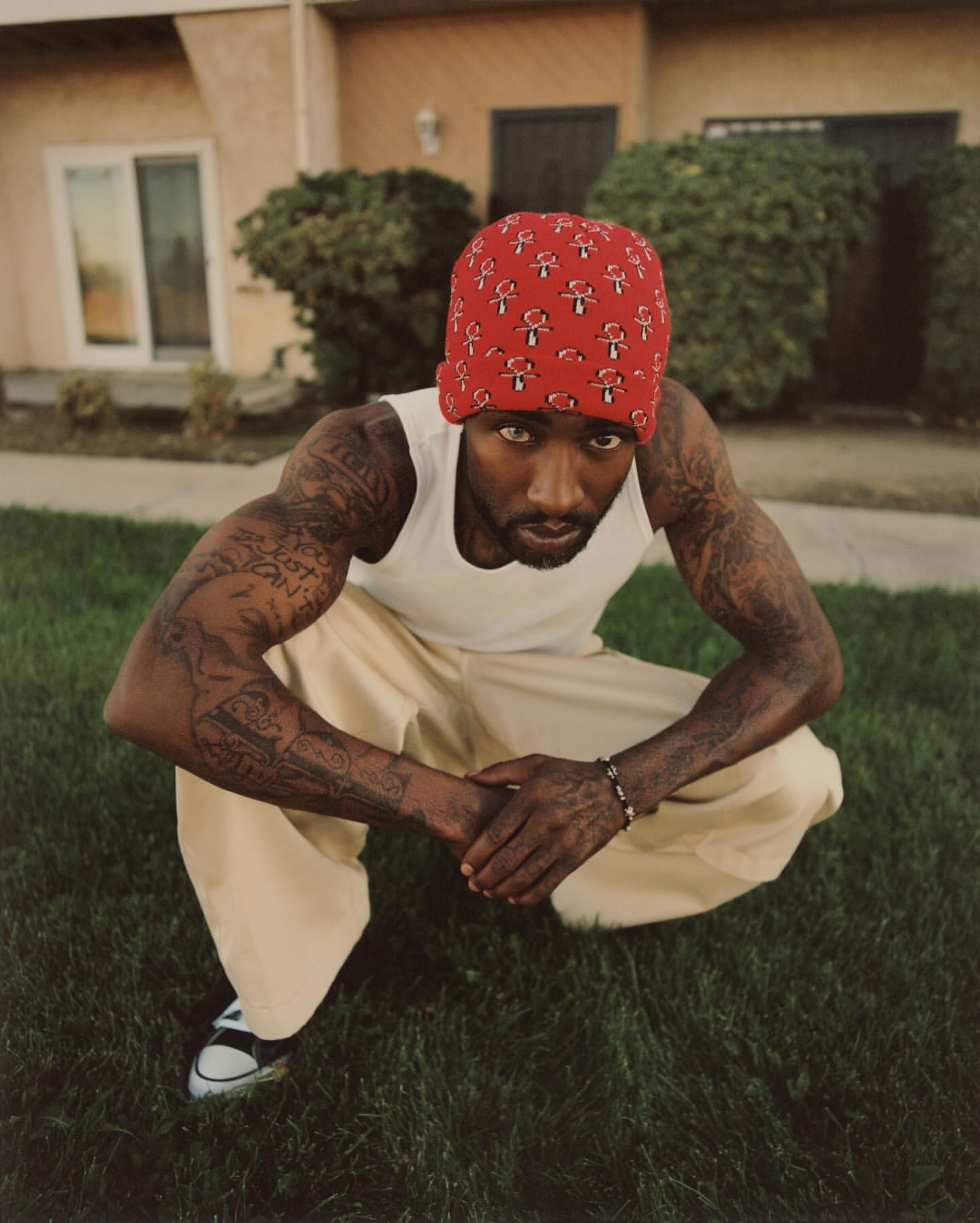 Man in tank top and bandana crouching on grass in front of a building with green shrubs, wearing black and white shoes.
