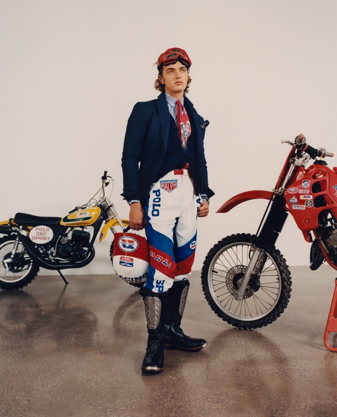 Male fashion model in racing attire poses indoors with two vintage motorcycles, one red and one yellow, creating a vintage racing aesthetic.