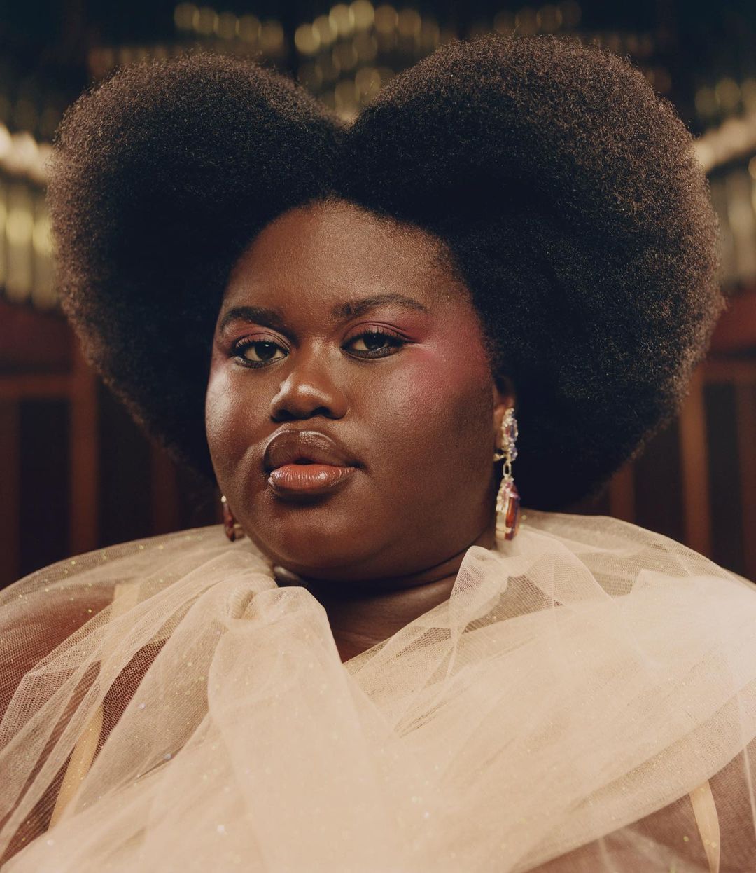 Close-up portrait of an adult woman with dark skin, large afro, and pink eye makeup wearing a tulle top and dangling earrings.