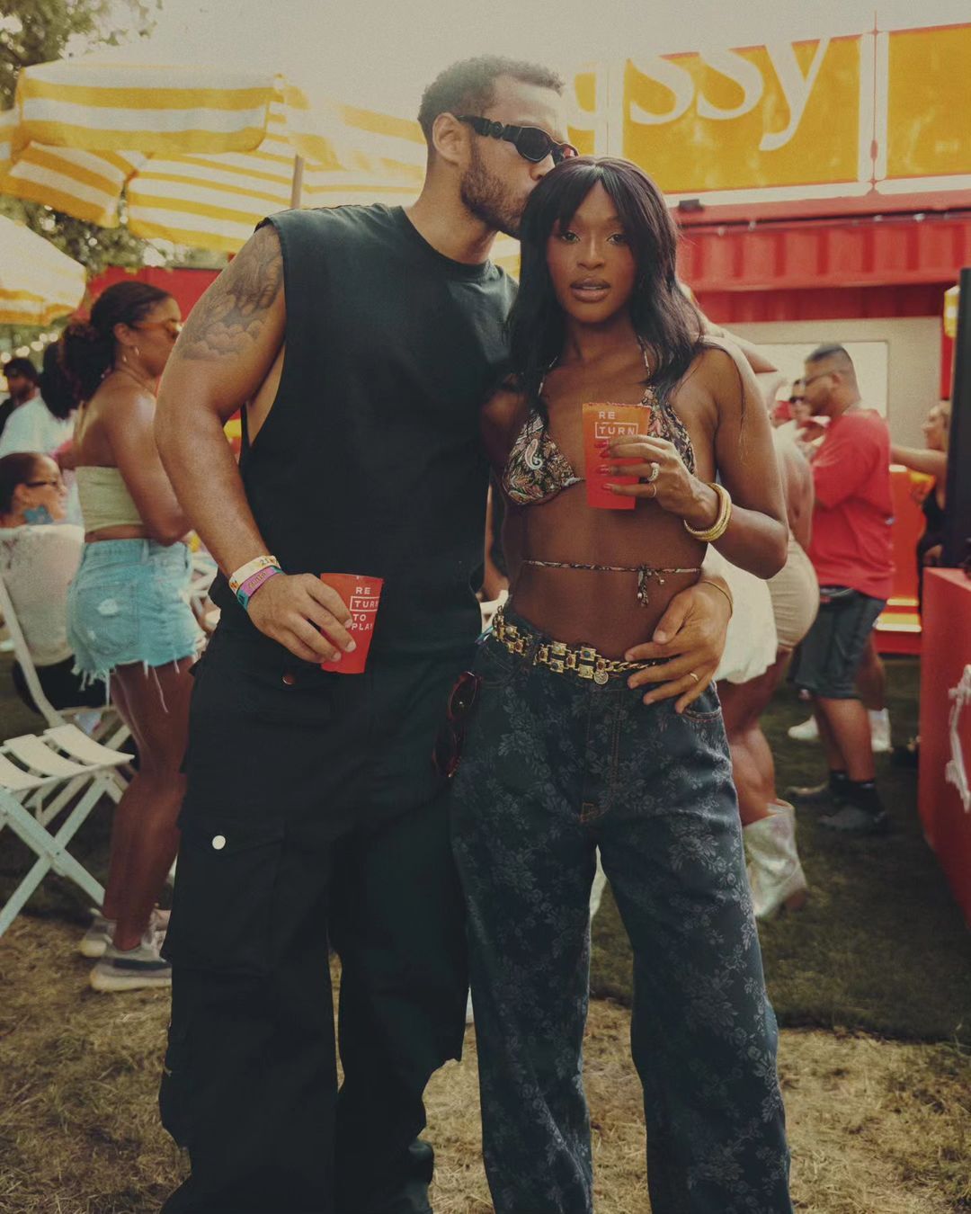 Outdoor portrait of a man kissing a woman on the head. Both are holding drinks at an outdoor event. Sunny weather with a crowd in background.