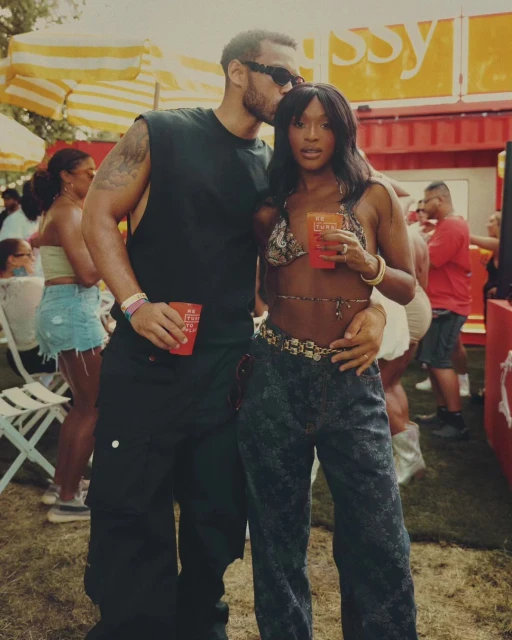 Outdoor portrait of a man kissing a woman on the head. Both are holding drinks at an outdoor event. Sunny weather with a crowd in background.