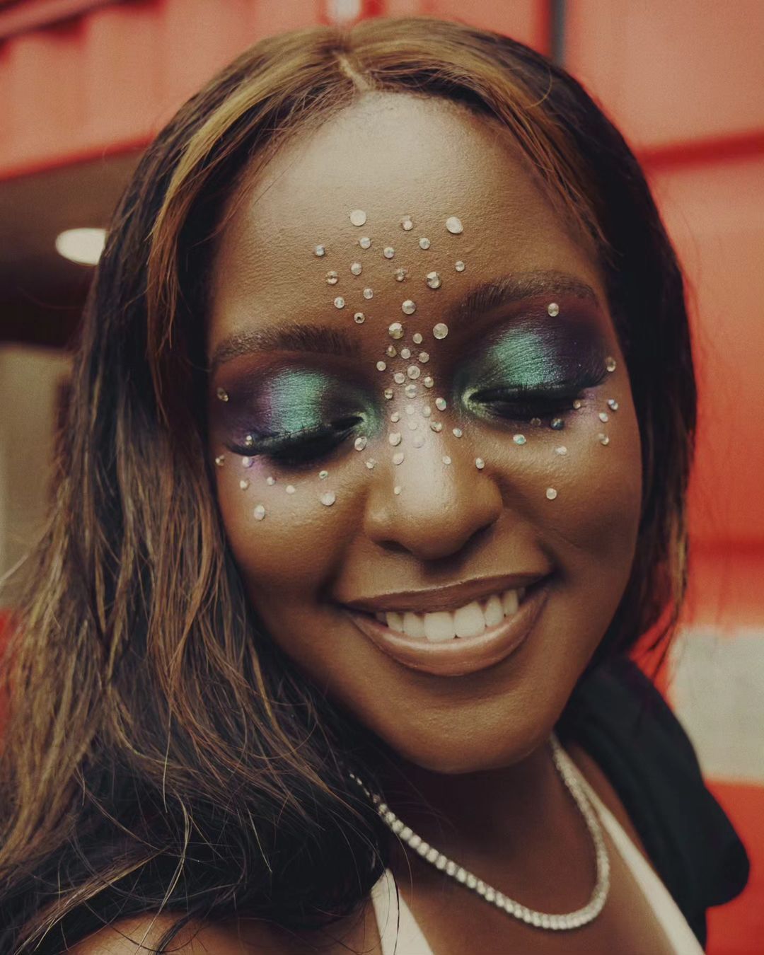 Close-up of a smiling adult female with brown hair wearing pearl necklace, colorful eye makeup, and facial gems against an orange wall.