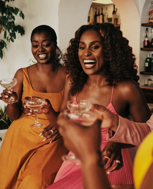 Three adult women dressed in formal dresses smiling and holding wine glasses at an indoor bar with bottles in the background.