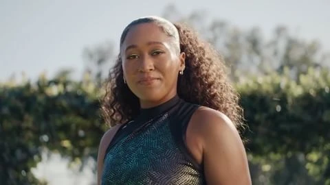 Portrait of Naomi Osaka wearing a patterned dark top outdoors with foliage in the background