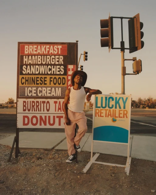 Young man in casual clothing and cowboy hat leans against a sign next to a food sign and traffic lights on a roadside under a clear sky.