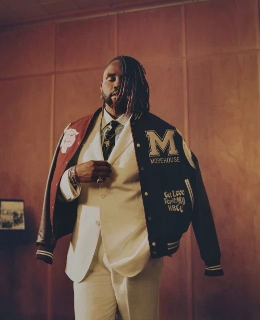 Portrait of a Black man in a cream-colored suit and letterman jacket, posing indoors against a reddish-brown wall with picture frame.