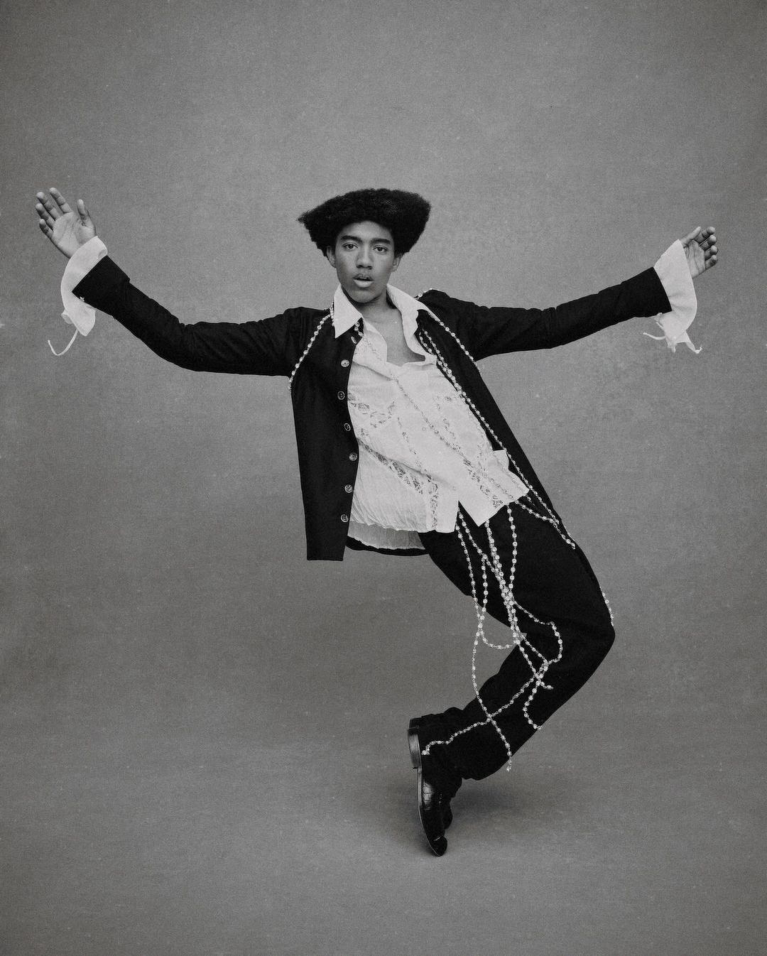 Monochromatic portrait of a stylish adult male in a suit jacket, lace shirt, and pearl strands, striking a dynamic pose in a studio.