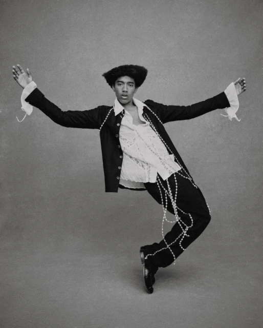 Monochromatic portrait of a stylish adult male in a suit jacket, lace shirt, and pearl strands, striking a dynamic pose in a studio.