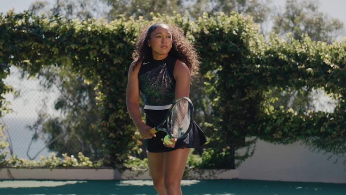 Female tennis player prepares to serve on a sunny outdoor court with a green ball, a black tennis outfit, and a racket.