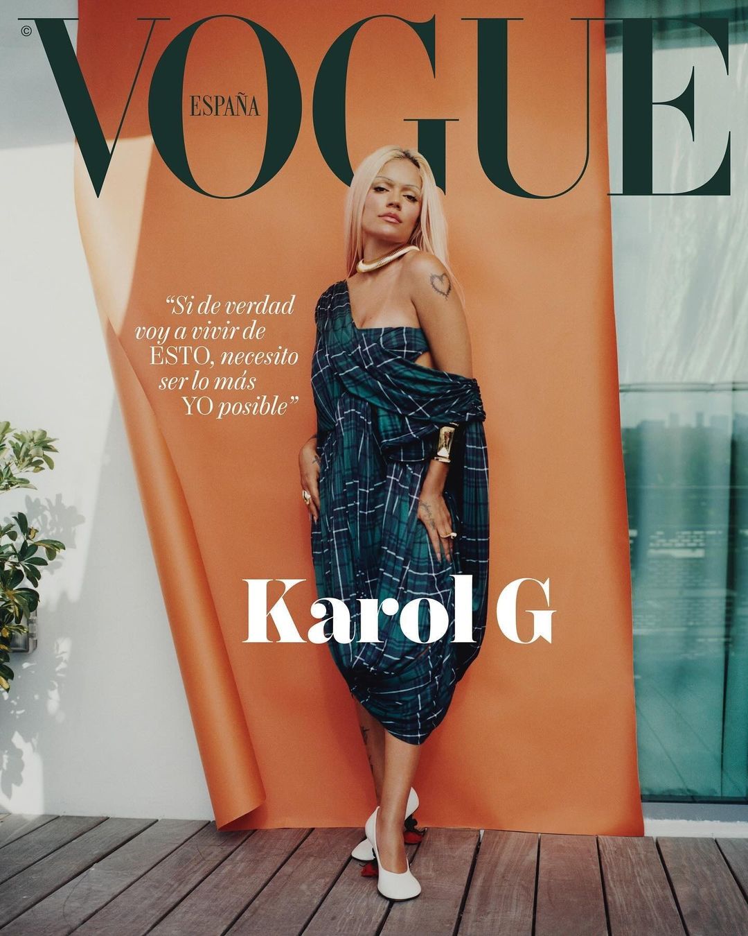 Karol G, photographed for Vogue España, poses in a green plaid dress with white heels, in front of an orange backdrop on a wood surface.