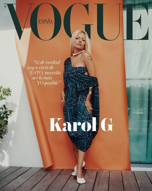 Karol G, photographed for Vogue España, poses in a green plaid dress with white heels, in front of an orange backdrop on a wood surface.