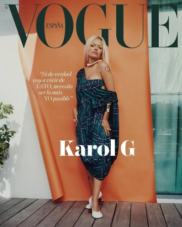 Karol G, photographed for Vogue España, poses in a green plaid dress with white heels, in front of an orange backdrop on a wood surface.