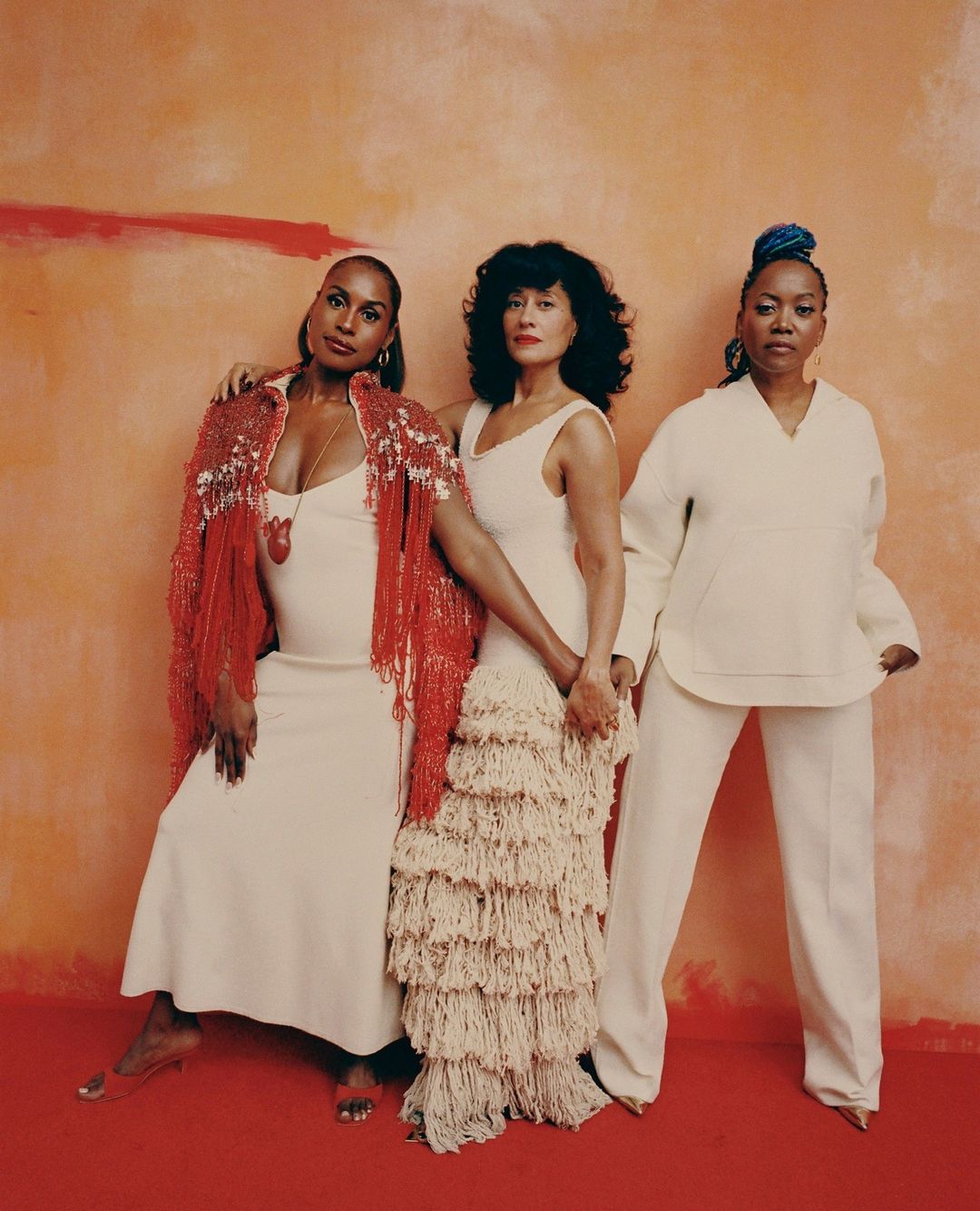 Three women pose in front of a textured orange background. They wear neutral-colored dresses and coordinating red and beige accents.