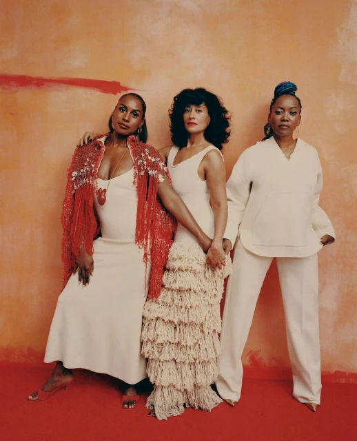 Three women pose in front of a textured orange background. They wear neutral-colored dresses and coordinating red and beige accents.