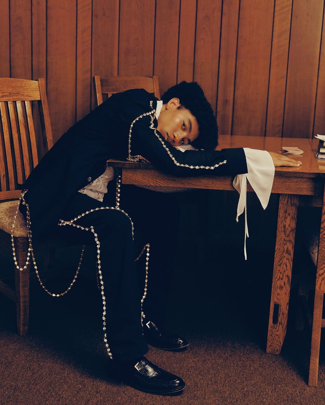 A young adult rests their head on a table wearing a suit jacket with beaded trim inside of a room with wood paneling.