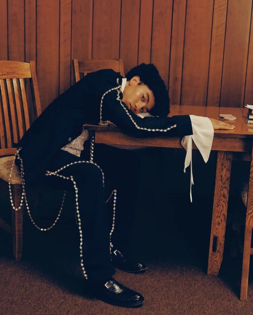 A young adult rests their head on a table wearing a suit jacket with beaded trim inside of a room with wood paneling.