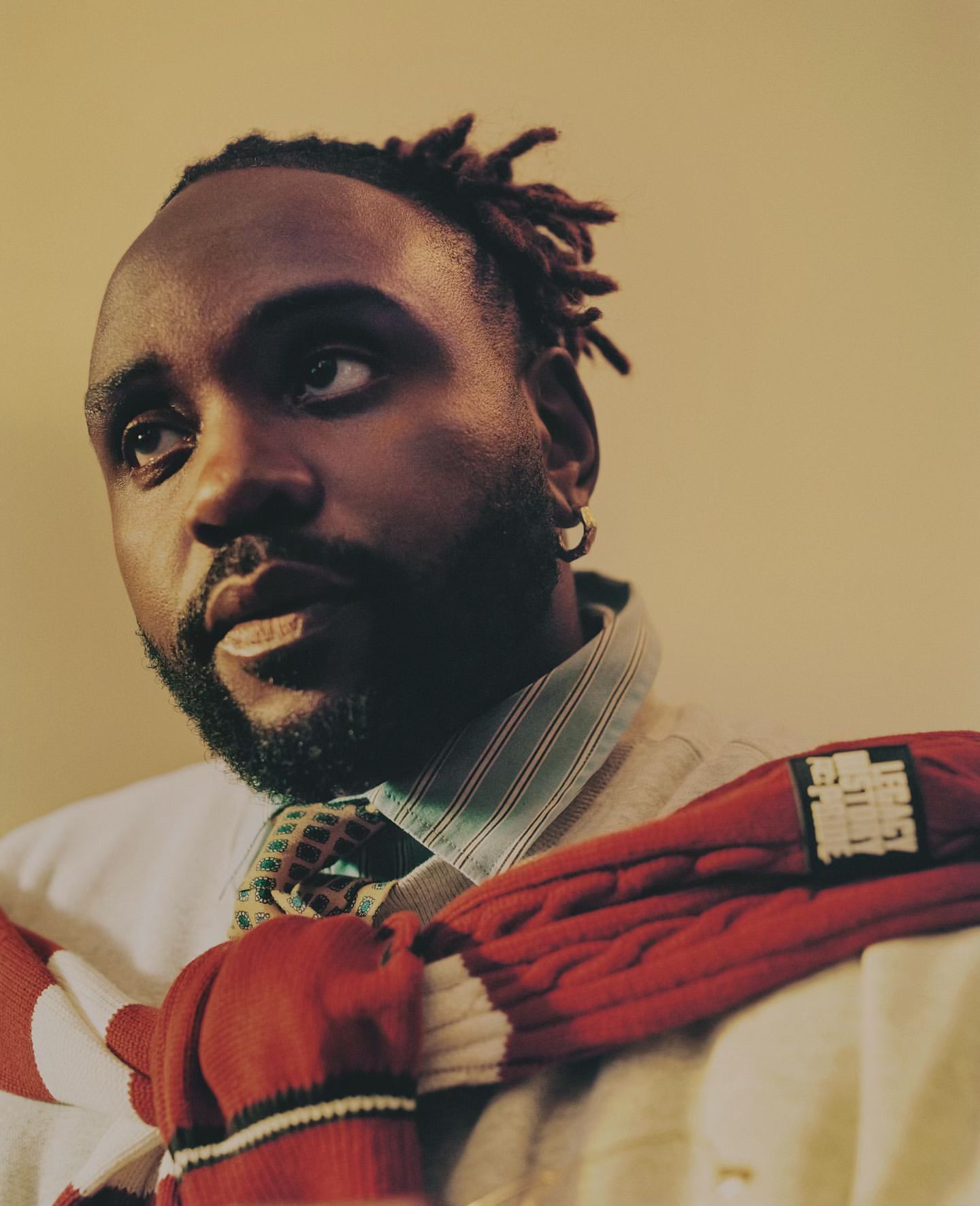 Portrait of a Black man with locs looking up, wearing a white button-down shirt, tie, and red sweater against a neutral background.