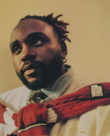Portrait of a Black man with locs looking up, wearing a white button-down shirt, tie, and red sweater against a neutral background.