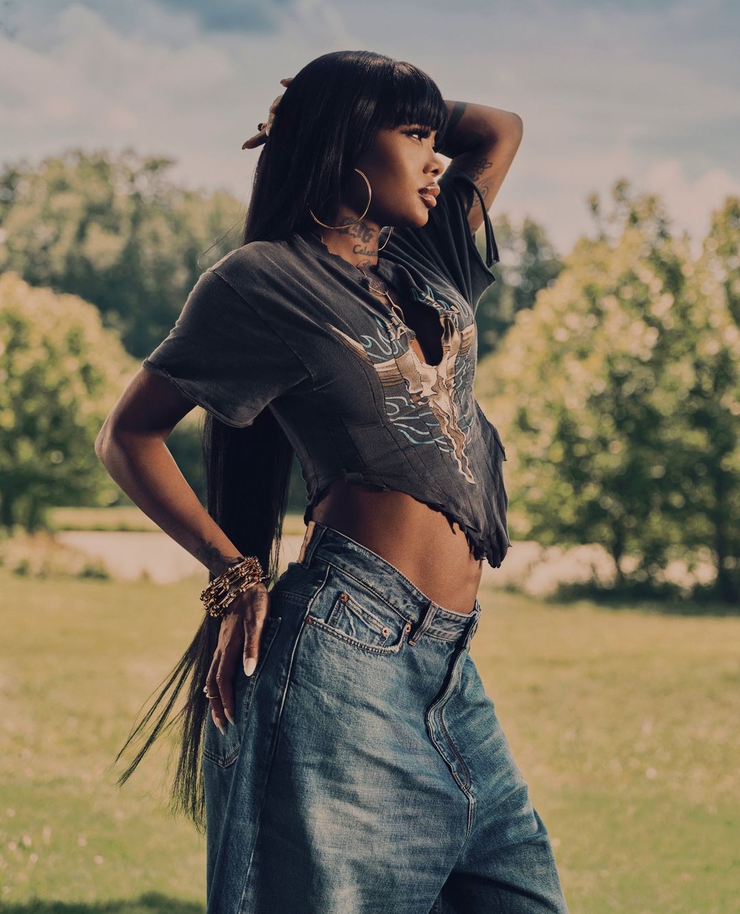 A woman with long dark hair poses in a t-shirt and baggy jeans against a backdrop of green grass and trees on an overcast day.