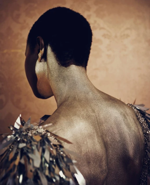 Back view of a person with dark hair and decorative silver shoulder pieces, set against a subtly patterned orange backdrop.