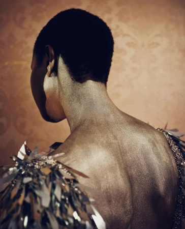 Back view of a person with dark hair and decorative silver shoulder pieces, set against a subtly patterned orange backdrop.