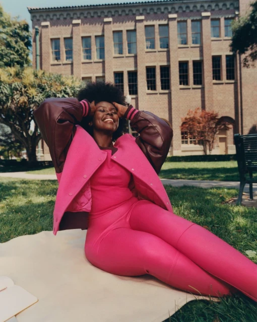 A smiling Black woman wearing a pink outfit and a varsity jacket is reclining on a blanket in a grassy area outside a brick building.