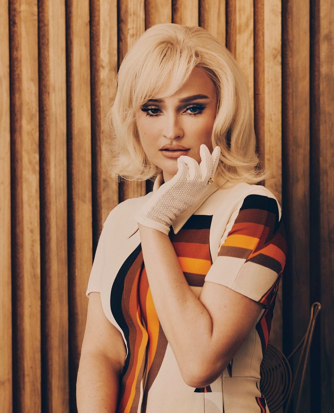 Blonde woman in retro dress poses against wood wall. She wears white gloves and has dark eye makeup in an indoor setting.