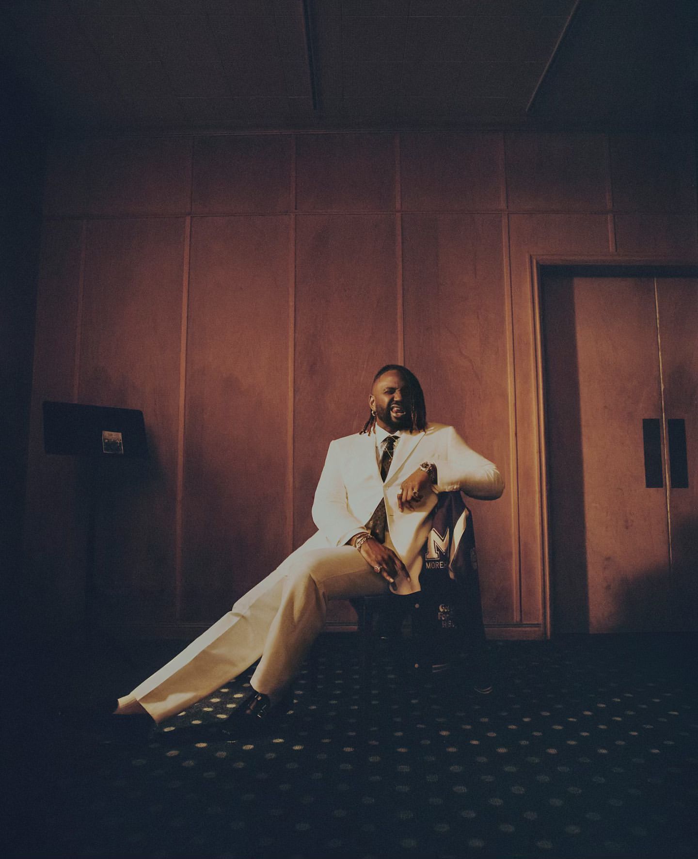 Portrait of an African American man in a white suit sitting indoors, posing against a paneled wall with a dark patterned floor.