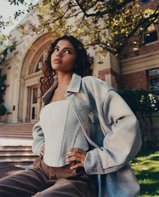 Portrait of a woman in a corset and jacket sitting outdoors near a brick building with trees, shot from a low angle.