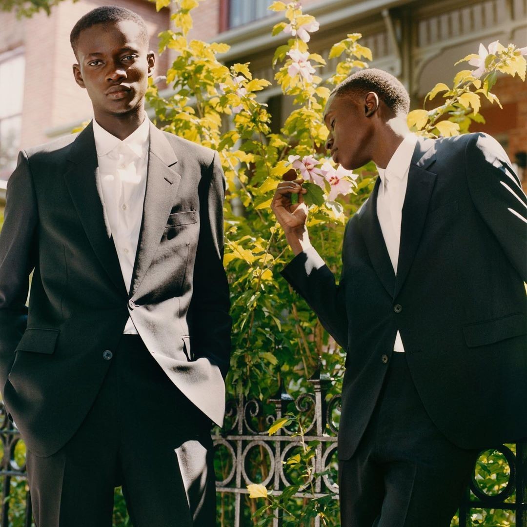 Two Black men in suits pose outdoors by foliage and an iron fence, one smelling a flower, showcasing fashion in natural light.