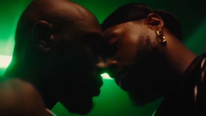 Close up of two Black men face to face in a dimly lit space. One man wears earrings and a headband with a green light illuminating the background.