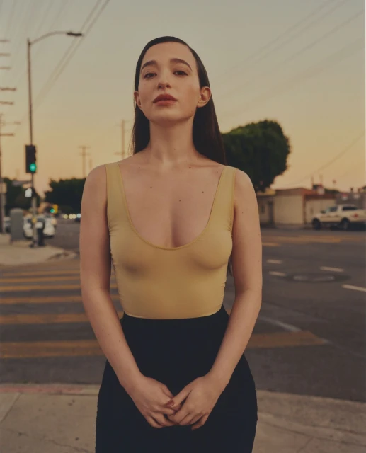Portrait of an adult woman in a beige leotard and black skirt standing on a crosswalk in an urban setting at dusk.