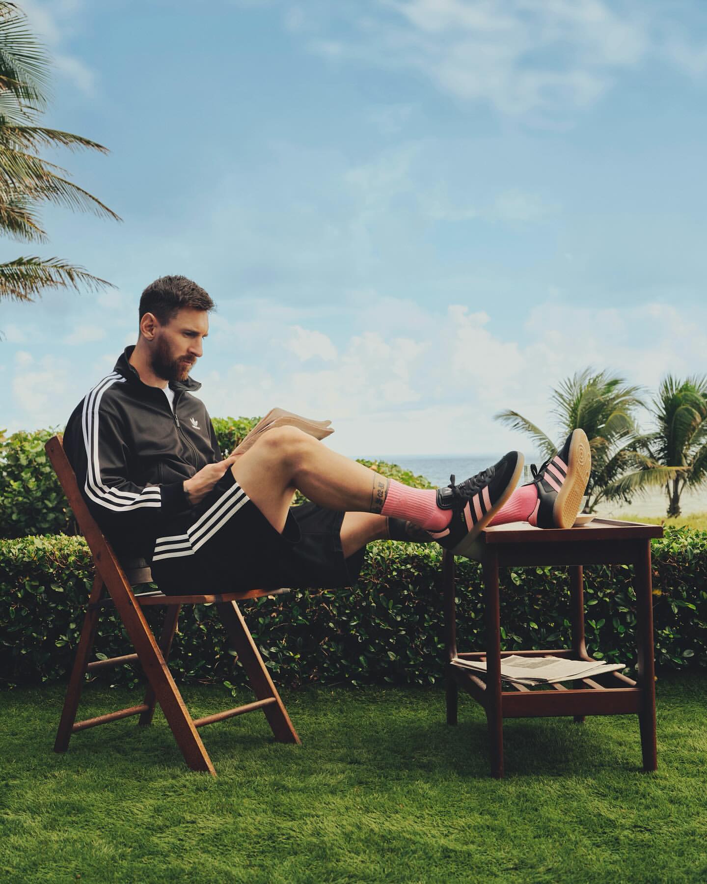 Lionel Messi relaxes on a wooden chair on green grass near a beach and palm trees, wearing an Adidas tracksuit and socks, reading a magazine.