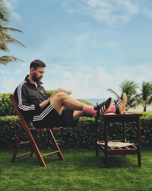 Lionel Messi relaxes on a wooden chair on green grass near a beach and palm trees, wearing an Adidas tracksuit and socks, reading a magazine.
