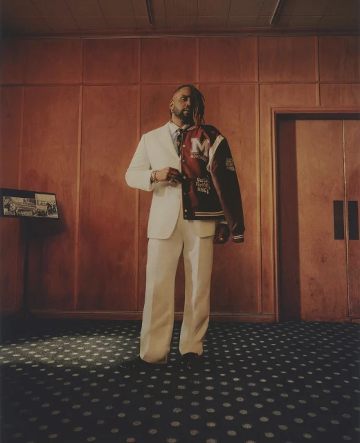 A stylish adult male in a white suit poses indoors with a letterman jacket, showcasing a blend of classic and modern fashion against a paneled wall.