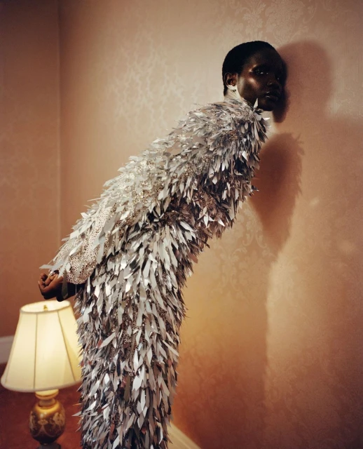 Fashion shot of an African American adult female wearing a full-length silver, leaf-like dress indoors, posing next to a lamp.