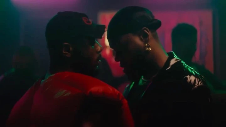Two African American men with dark hats face each other in a dimly lit room with red and green lighting, creating a dramatic and intimate atmosphere.