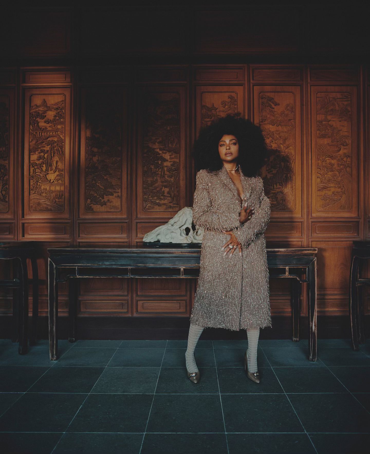 A fashionable Black woman with an afro poses in a beaded coat and tights in an interior with ornate wooden panels.