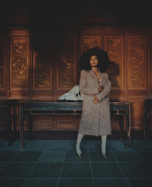 A fashionable Black woman with an afro poses in a beaded coat and tights in an interior with ornate wooden panels.
