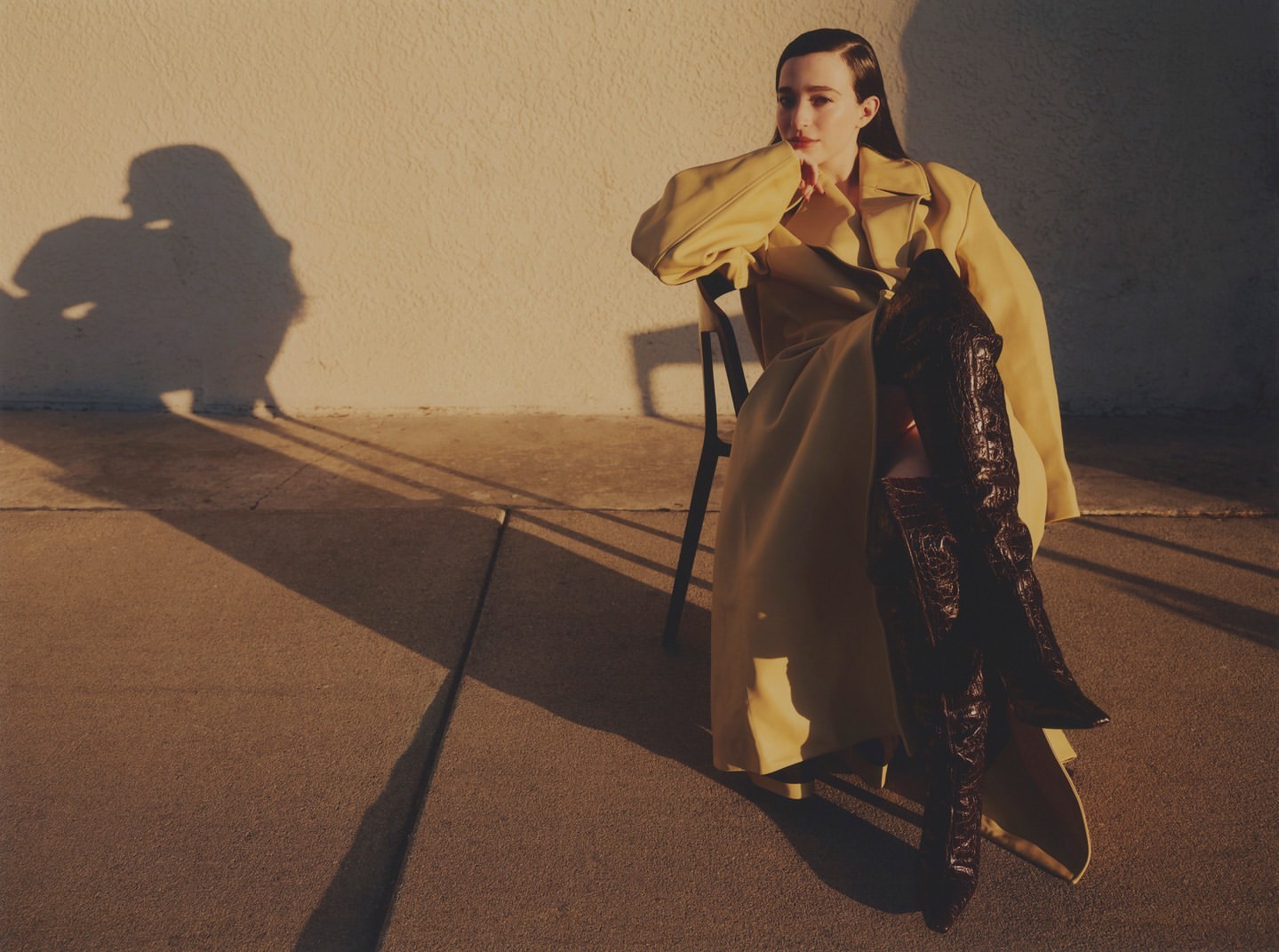 A stylish woman in a yellow trench coat poses while seated on a chair, accentuating the eye-catching brown knee-high boots in outdoor lighting.