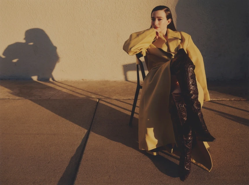 A stylish woman in a yellow trench coat poses while seated on a chair, accentuating the eye-catching brown knee-high boots in outdoor lighting.