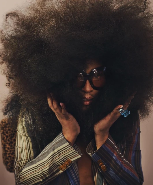 Striking portrait of an African American person with a large Afro, wearing dark glasses and a striped jacket with a leopard-print bag.