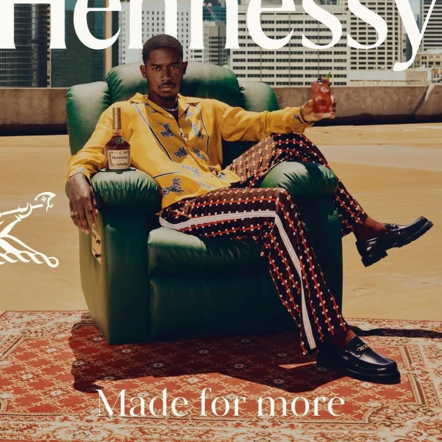 Stylish man in patterned shirt and pants sits in a green armchair, holding a cocktail, with a Hennessy bottle visible and city backdrop.