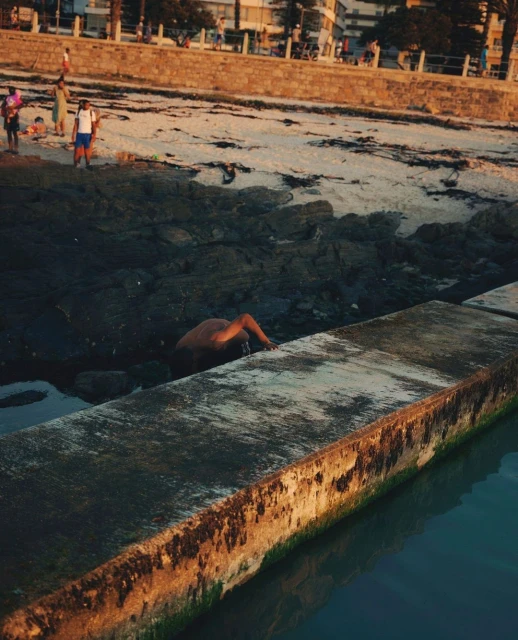 Adult dips in a rock pool at a sandy beach near buildings; warm sunlight on concrete wall and water reflections visible.