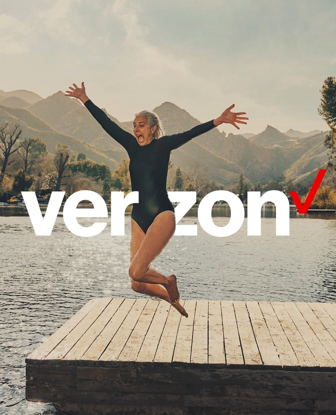 A woman in a black swimsuit jumps with arms raised from a wooden dock into a lake, with mountains visible in the background.