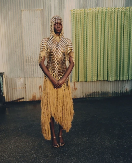 A dark-skinned fashion model wearing a gold dress with beaded hood stands in front of a corrugated metal wall and patterned green curtain.