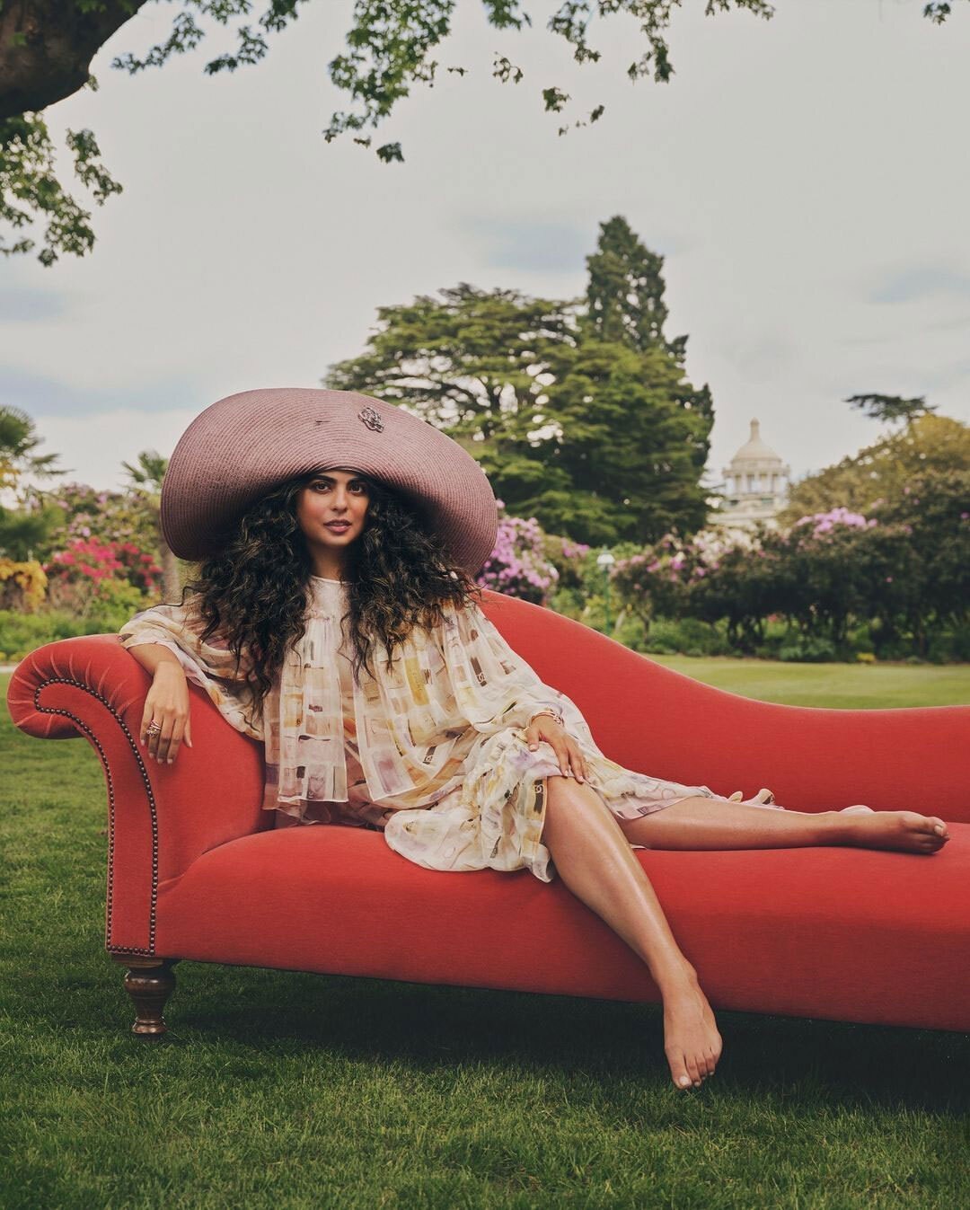 A woman in a patterned dress and large hat reclines on a red chaise lounge in a garden with trees and a building in the background.