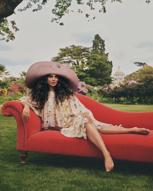 A woman in a patterned dress and large hat reclines on a red chaise lounge in a garden with trees and a building in the background.