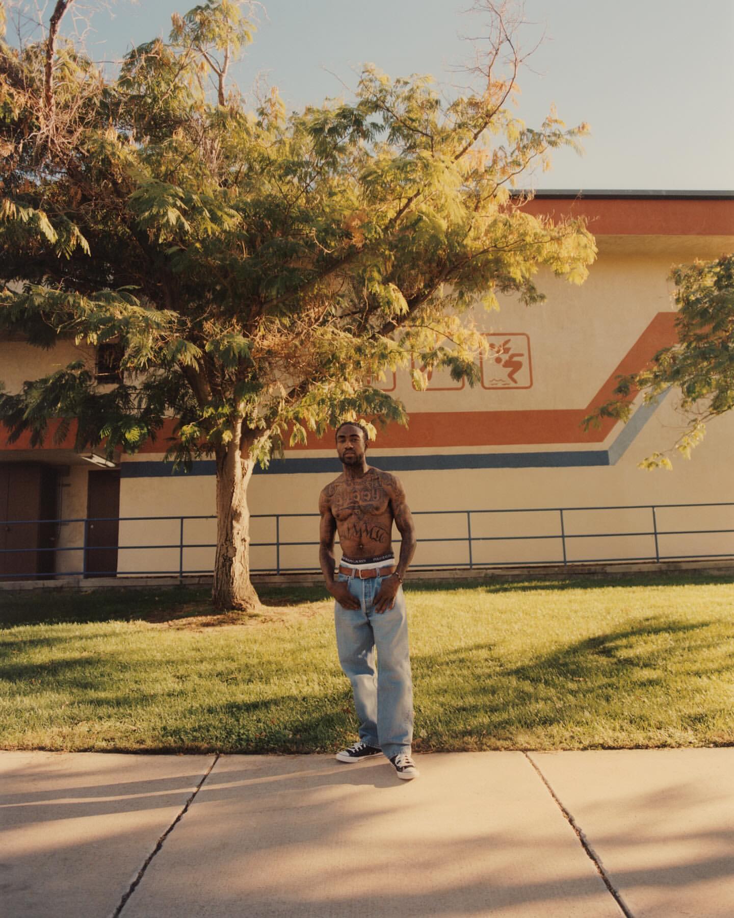 A shirtless man with tattoos stands on a sidewalk near a building and a green lawn with a large tree in the background outdoors.