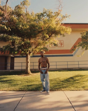 A shirtless man with tattoos stands on a sidewalk near a building and a green lawn with a large tree in the background outdoors.